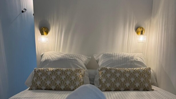 Cama com lençóis brancos e almofadas estampadas, duas luminárias fixadas na parede e janela com cortinas translúcidas.