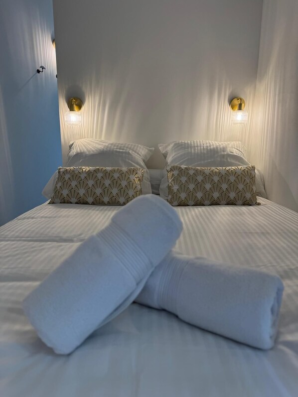 Een bed met witte lakens en gestreepte kussens, twee wandlampen en een raam met dunne gordijnen.