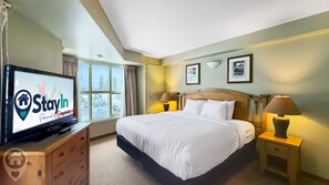 A hotel room with a bed, bedside lamps, a television displaying the StayIn logo, a window with a view of a snowy parking lot, and framed pictures on the wall. - Polaris Lodge / Ski-In/Ski-Out / Sleeps 10 (Kimberley)
