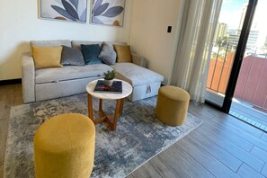 A modern living room with a grey sofa, two round ottomans, a small table with a plant, and a city view through a sliding door. - EON Zone 10. Luxury, views, and comfort—all in one apartment. (Ciudad de Guatemala)