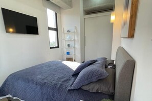 Ein Schlafzimmer mit einem Bett, einem an der Wand montierten Fernseher, einem Fenster und einem weißen Kleiderschrank.