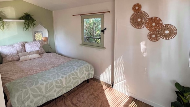 A bedroom with a bed, a window, a wall-mounted light fixture, and a plant.