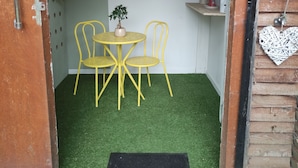 A small room with a green artificial turf floor, a round table with two chairs, a potted plant, and a small shelf with a lamp and a cup.