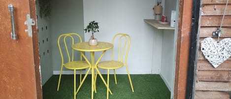 Una stanza piccola con pavimento in erba sintetica verde, un tavolo rotondo con due sedie, una pianta in vaso e un piccolo ripiano con una lampada e una tazza.