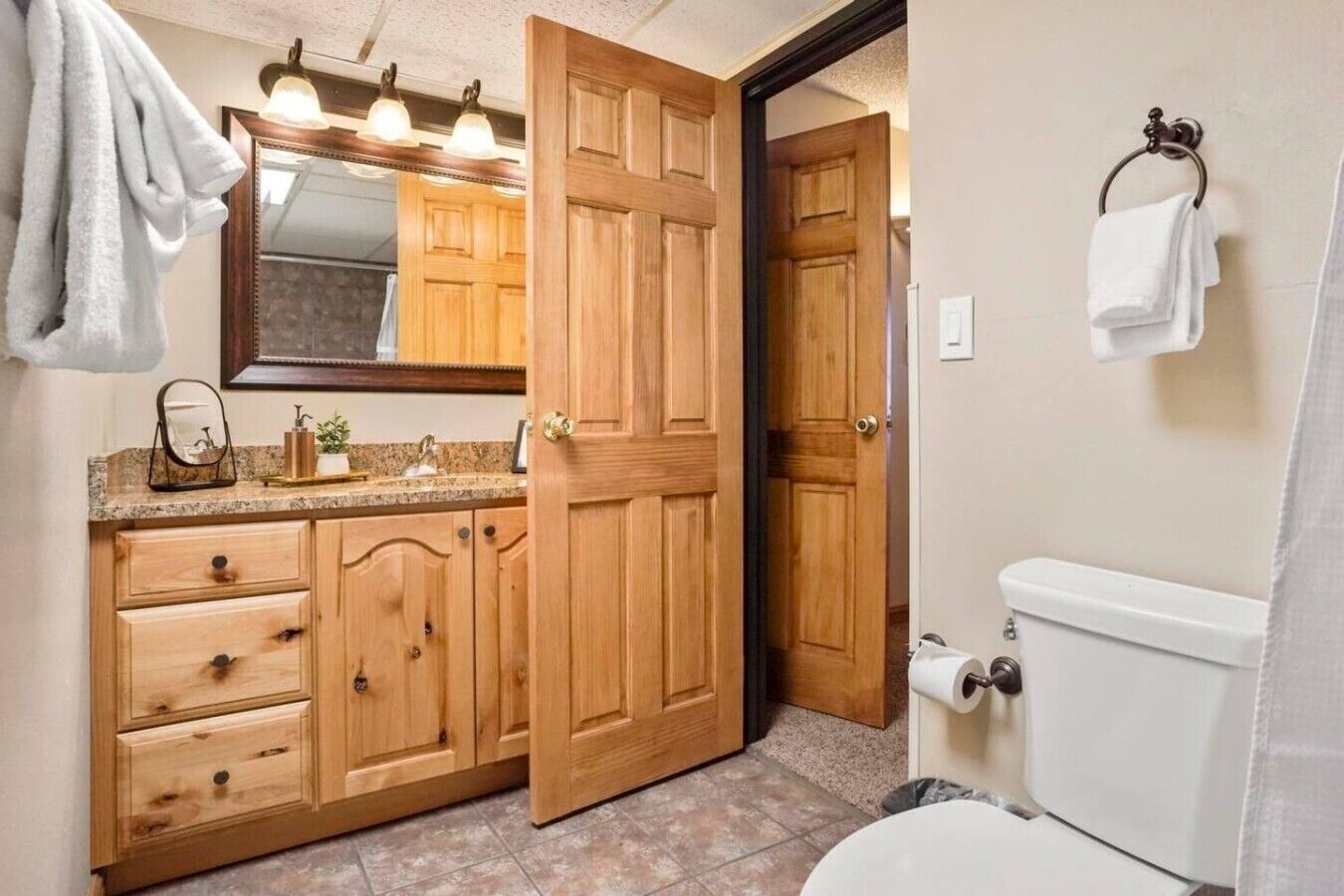 A bathroom with wooden cabinetry, a toilet, and a mirror with lights above the sink.