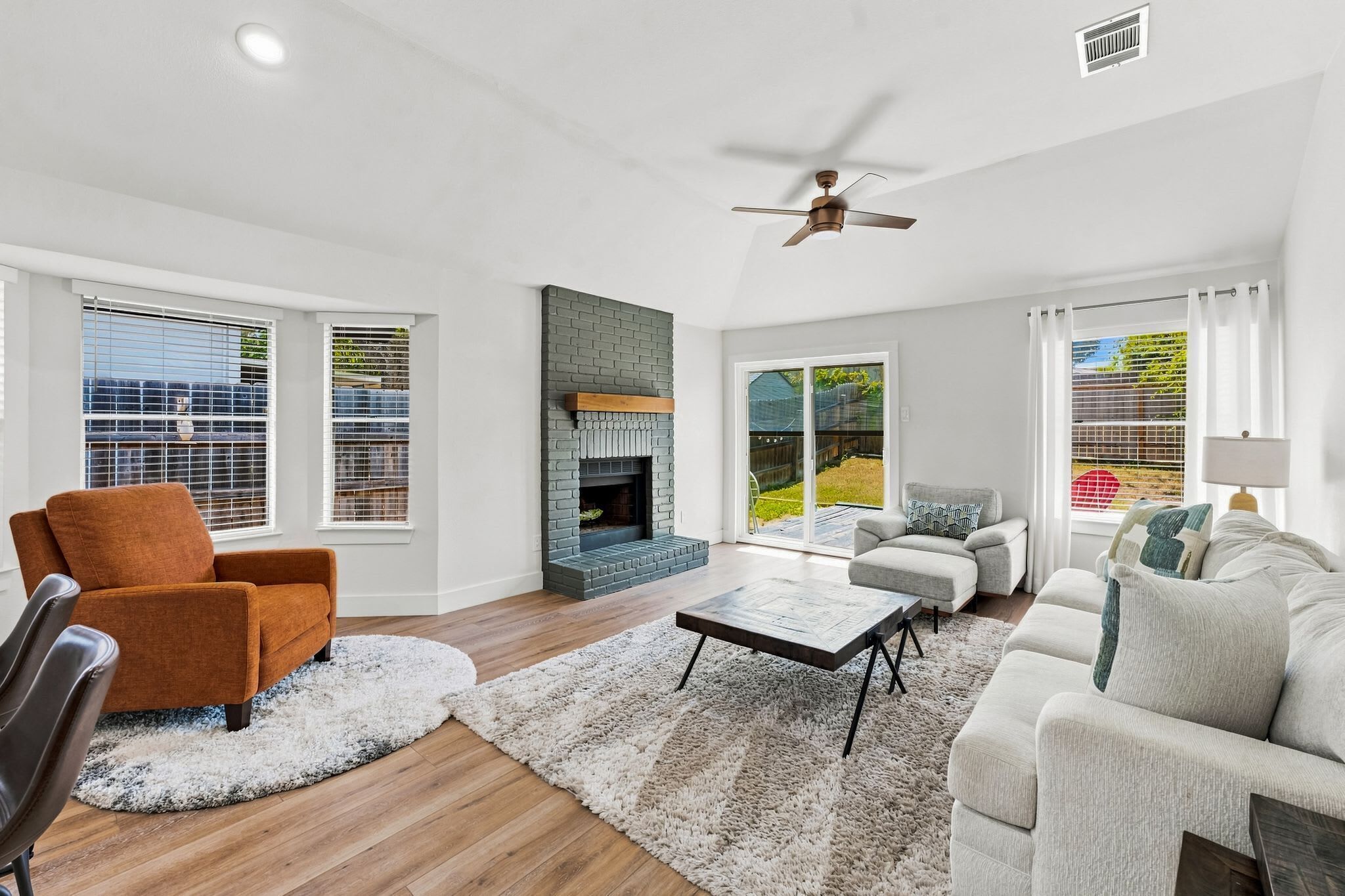A spacious living room with a fireplace, a ceiling fan, and large windows.
