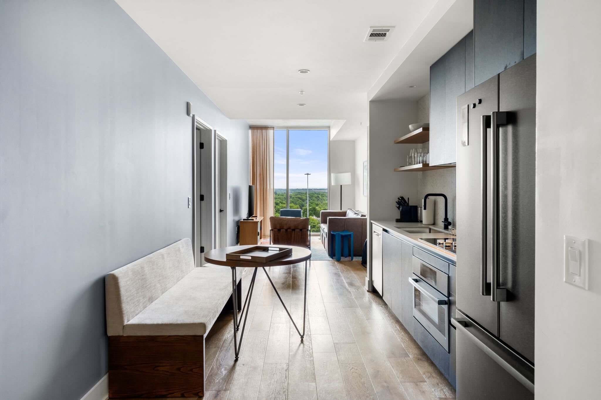 A modern kitchen with a large refrigerator, a dining area with a bench and table, and a view of greenery outside.