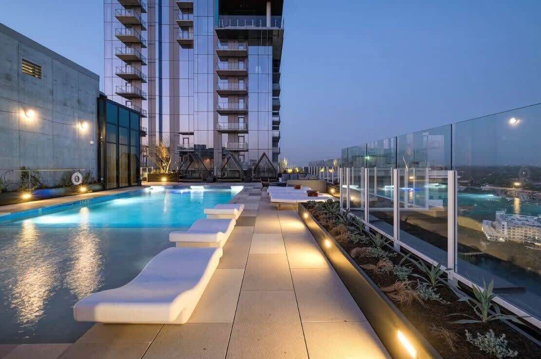 A rooftop pool with tiered seating and a glass balustrade, surrounded by a high-rise building with balconies.