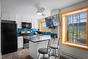 A modern kitchen with a central island, bar stools, a flat-screen TV, and a large window overlooking the outdoors. - Village Peak 9 Inn Ski-in/Ski-out Walk to Main St (Breckenridge)