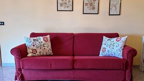 Un canapé rouge orné de coussins à motifs floraux, dans une pièce dont le sol est recouvert de carreaux et où l’on peut voir, au mur, des œuvres d’art florales encadrées.