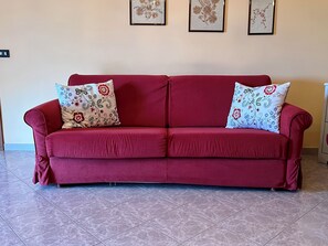 Un sofá rojo con cojines de estampado floral en una habitación con suelo de baldosas y cuadros de motivos florales en la pared.