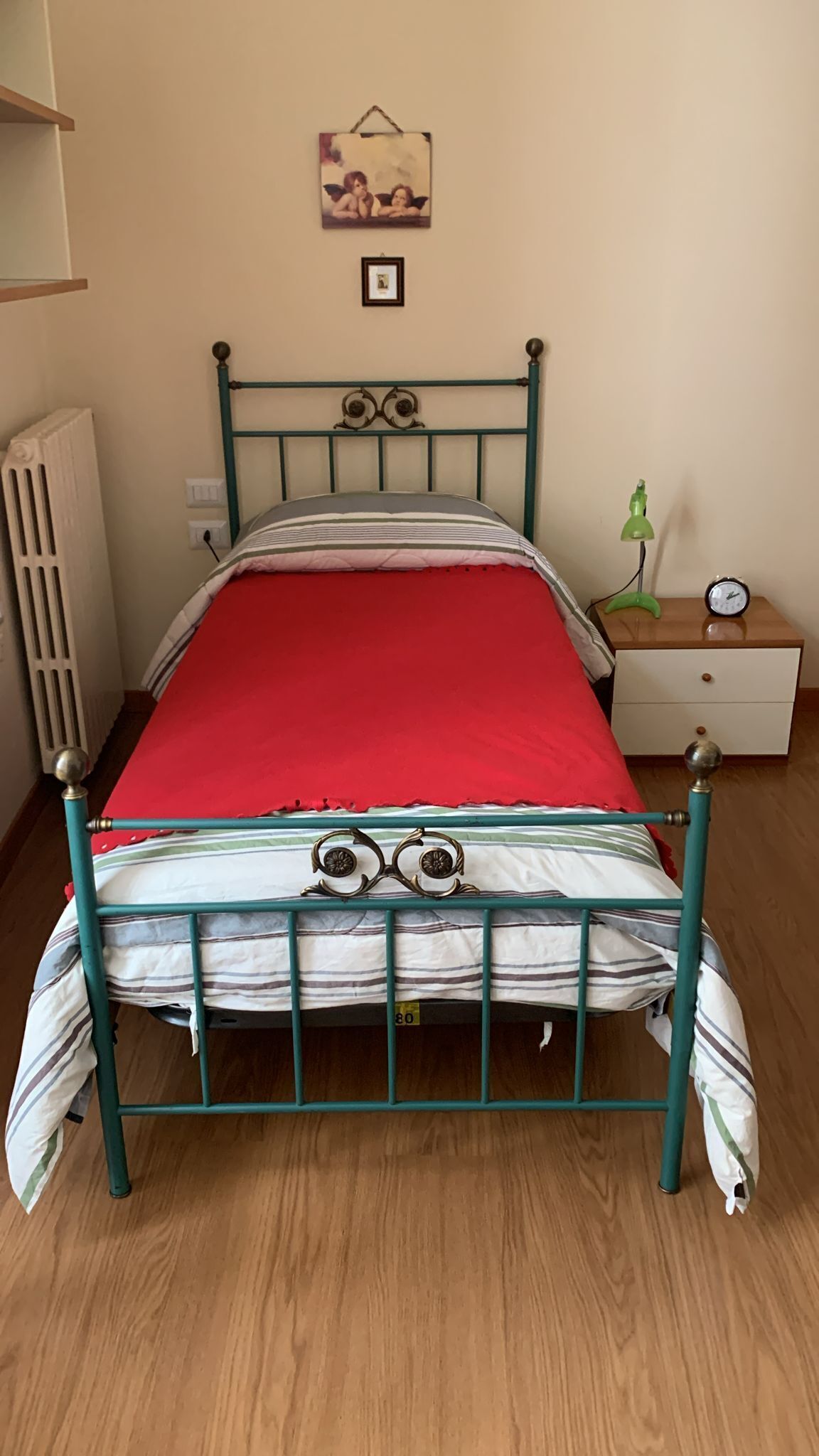 Een eenpersoonsbed met een rode matras, metalen frame en een hoofdbord met decoratieve details. Rechts staat een nachtkastje met een klok en een lamp.