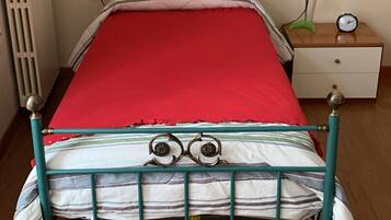 Een eenpersoonsbed met een rode matras, metalen frame en een hoofdbord met decoratieve details. Rechts staat een nachtkastje met een klok en een lamp.