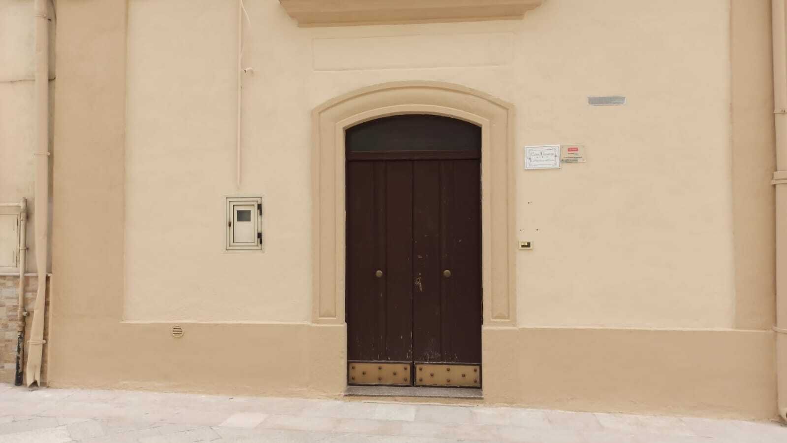 Facciata di un edificio con una porta in legno scuro a due battenti, una piccola finestra rettangolare e una targa sulla parete.