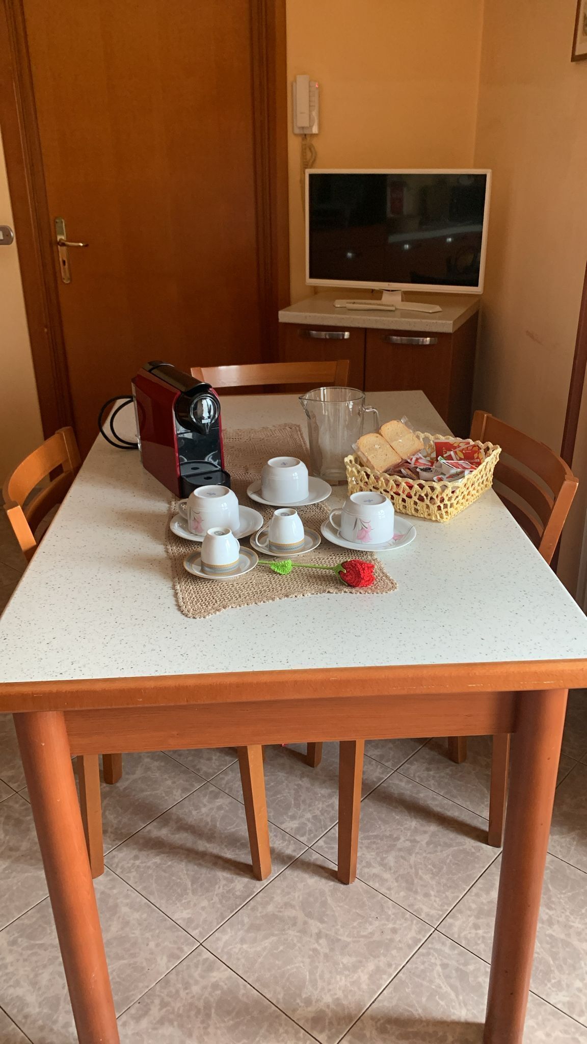 Un tavolo da pranzo con una macchina da caffè, tazze, piattini, un boccale di vetro e un cesto di pane.