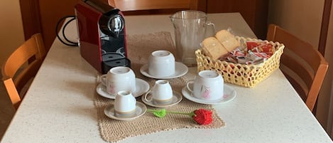 A dining table with a coffee maker, cups, saucers, a glass pitcher, and a basket of bread.