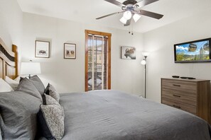 Ein Schlafzimmer mit einem Bett, einem Deckenventilator, einem Fernseher, einer Kommode und einem Fenster mit Jalousien.