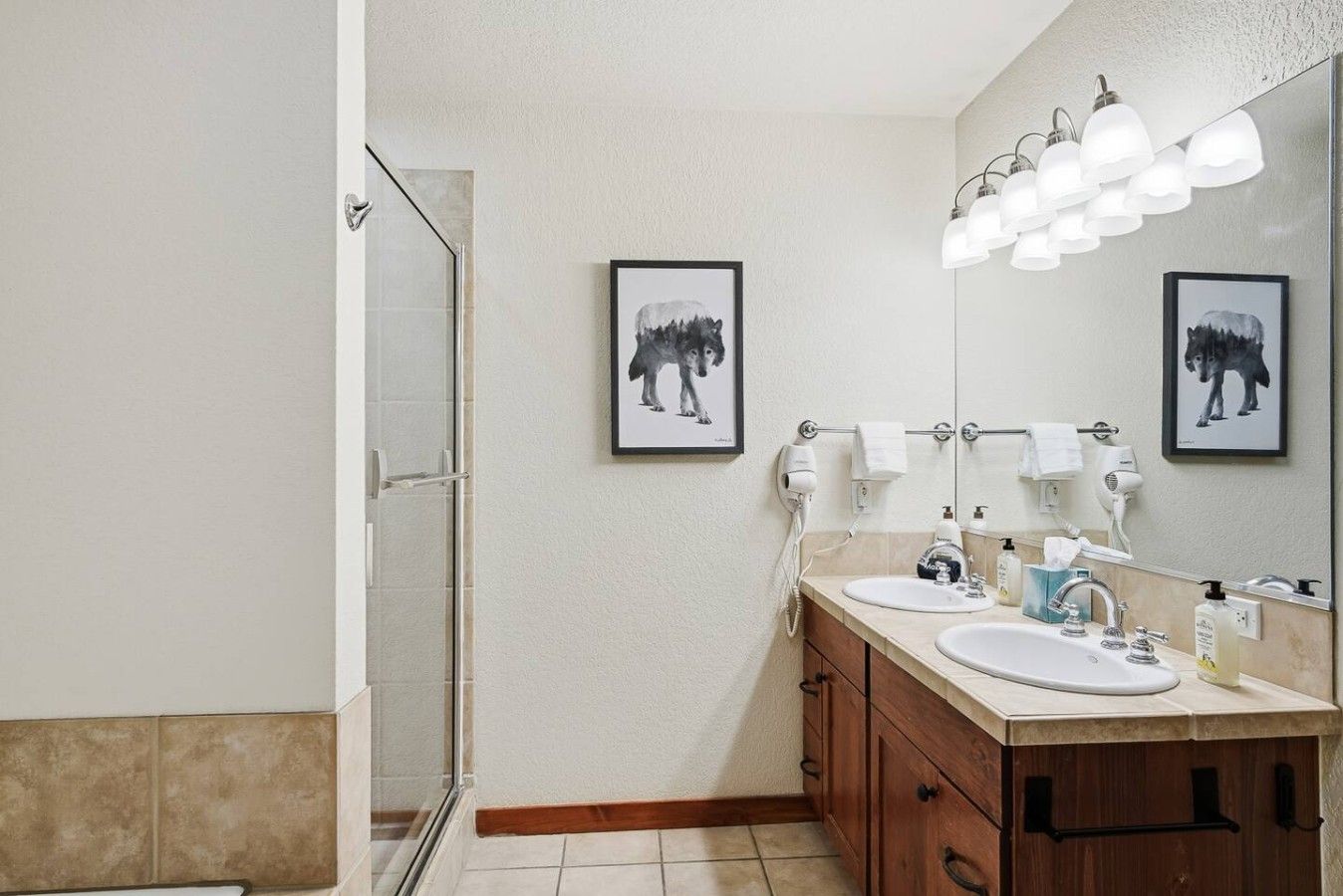 A bathroom with a shower, two sinks, a mirror, and framed pictures on the wall.
