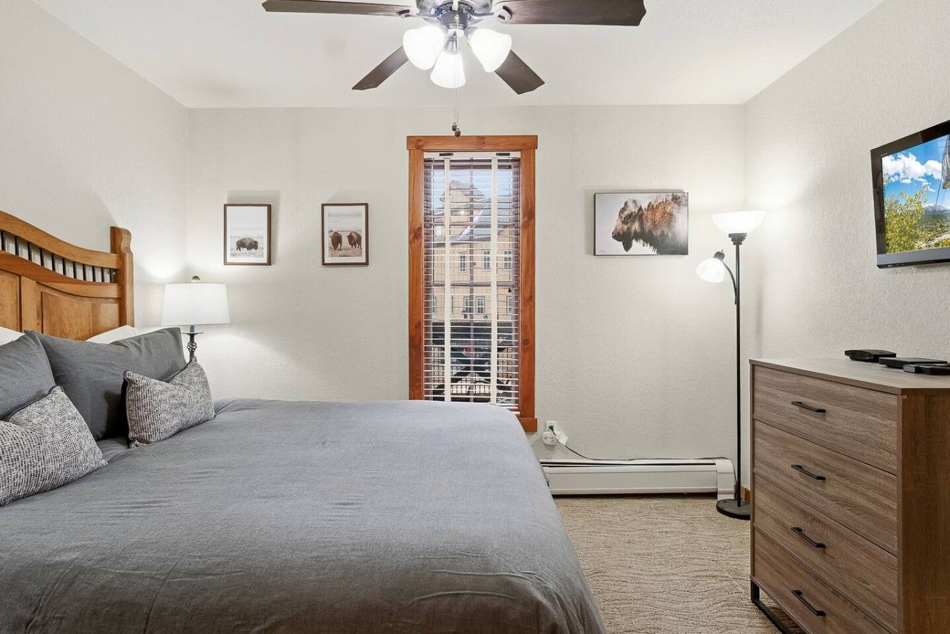 A bedroom with a bed, a nightstand, a dresser, a ceiling fan, a window with blinds, and wall-mounted artwork.