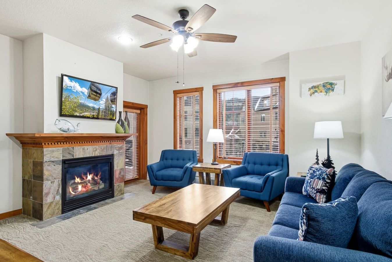 A living room with a fireplace, two blue armchairs, a wooden coffee table, and a ceiling fan.