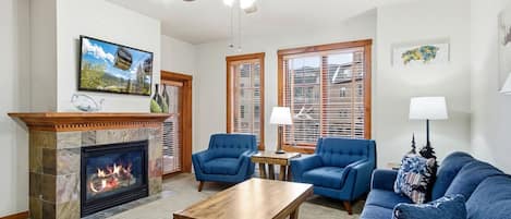 A living room with a fireplace, two blue armchairs, a wooden coffee table, and a ceiling fan.