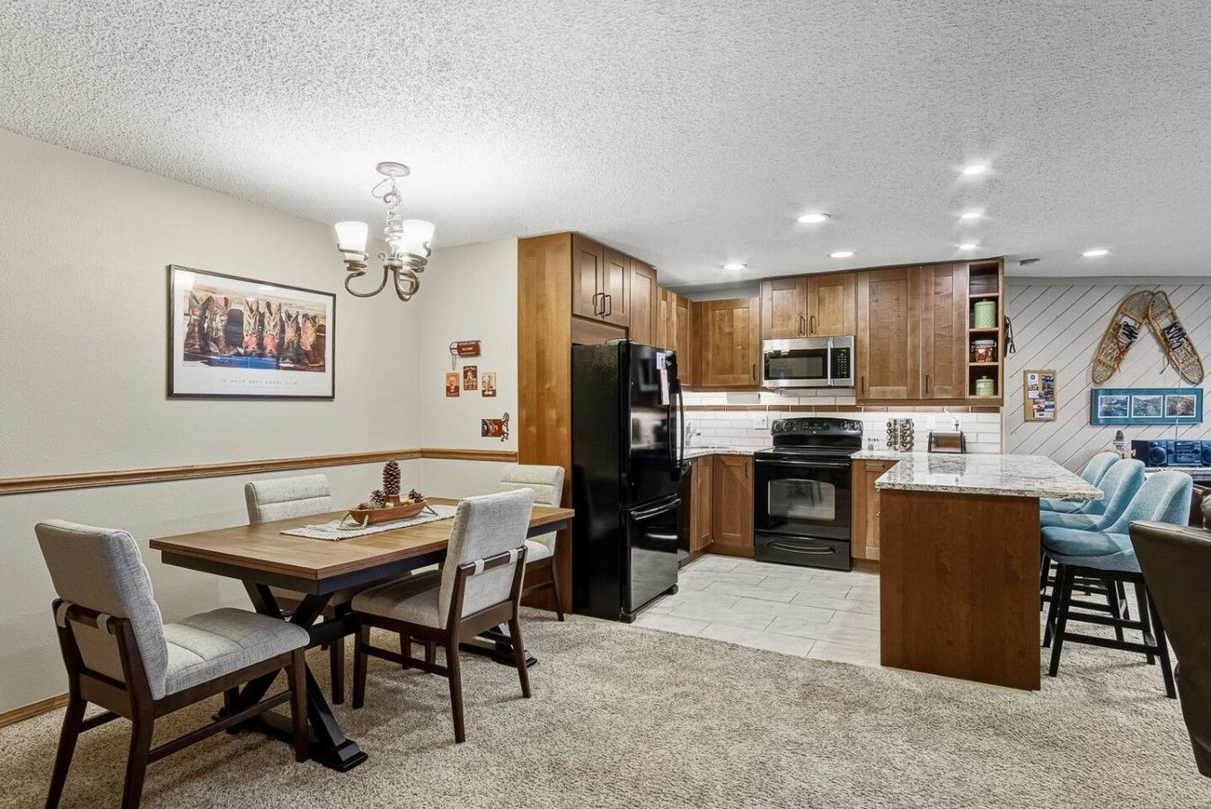 A modern kitchen with wooden cabinets, a black refrigerator, a microwave, and a stove. There is a dining area with a table and chairs.