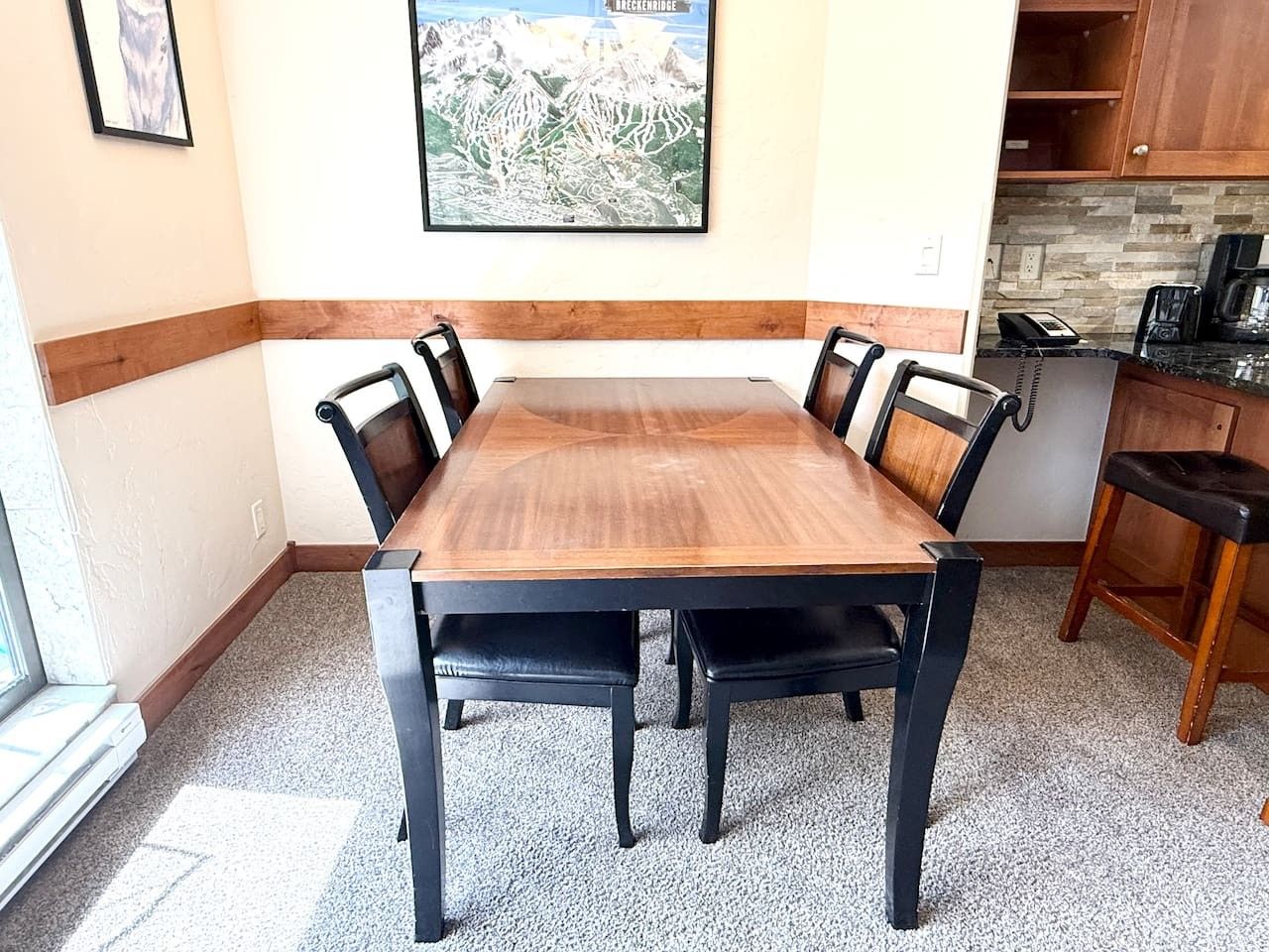 A dining area with a wooden table and black chairs, a kitchen area with a coffee maker, and a framed picture on the wall.