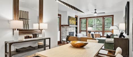 A modern living room with a dining area, large windows, a fireplace, and a ceiling fan.
