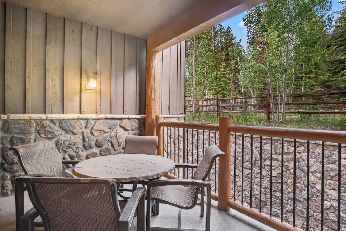 A covered outdoor seating area with stone walls, a wooden railing, and a view of trees.
