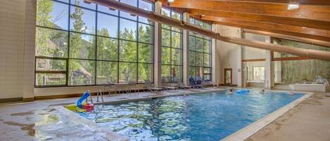An indoor swimming pool with clear glass walls, wooden beams, and a view of trees outside.