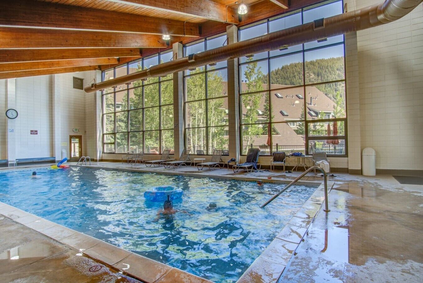 An indoor swimming pool with clear windows, lounge chairs, and a visible clock on the wall.