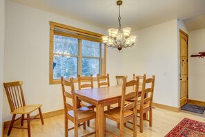 Ein Esszimmer mit Holztisch und -stühlen, einer Kristallleuchter, einem großen Fenster mit Blick auf Bäume und einer Holztür.