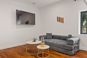 Un salon moderne avec un canapé gris, deux tables rondes, un téléviseur fixé au mur et une fenêtre donnant sur un beau paysage.