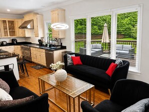 A modern kitchen with wooden cabinets, a dining area with a black sofa, and a view of a green garden through large windows. - Jamestown 4BR Estate (Jamestown)