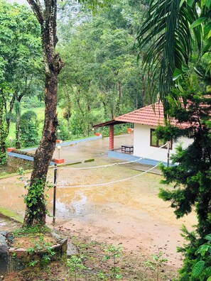 Un espacio al aire libre techado con una red de voleibol, rodeado de abundante follaje y un pequeño edificio con techo rojo.