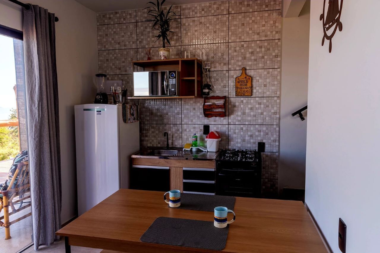 A compact kitchen with a refrigerator, microwave, and stove. There is a dining table with two mugs and a bench. A potted plant is on the wall, and a sign is visible.