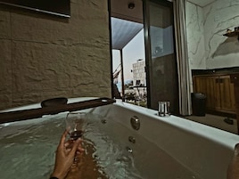Una persona relajándose en un jacuzzi con una copa de vino, una ventana y vistas a edificios.