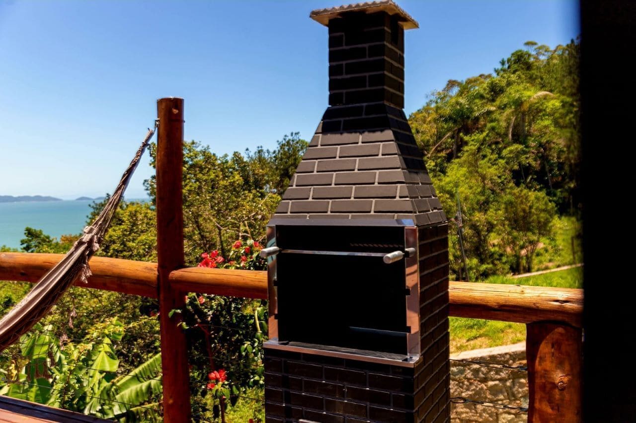 Churrasqueira de tijolos com chaminé, emoldurada por vegetação exuberante e céu azul límpido.
