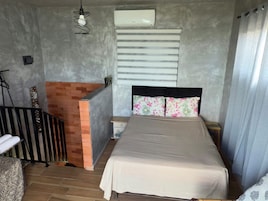 Un dormitorio con cama, mesita de noche, una pequeña mesa, un tabique de ladrillo, un radiador y una ventana con persianas.