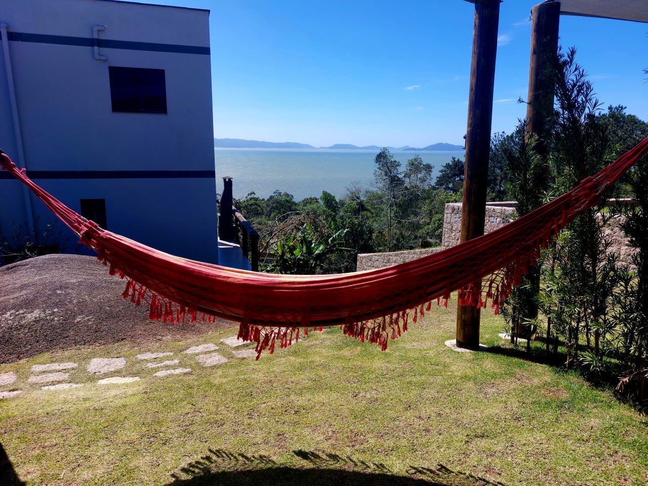 Una hamaca atada entre dos postes de madera en un espacio al aire libre con césped, con vista a un cuerpo de agua y colinas al fondo.