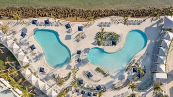 An aerial view of a resort with two large swimming pools, sun loungers, and palm trees.
