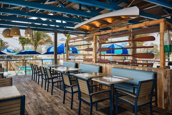 An outdoor dining area with wooden tables and chairs, a poolside view, and blue umbrellas.