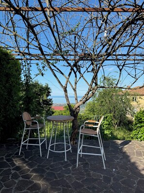A patio with two white metal chairs and a table, under a tree with a view of the sea. - Stylish, high-end villa in the hills of Versilia with ocean views (Toscana)