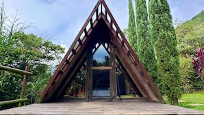 A wooden structure with a triangular roof and a glass door, surrounded by greenery and a clear sky. - All Natural Glamping (Copacabana)