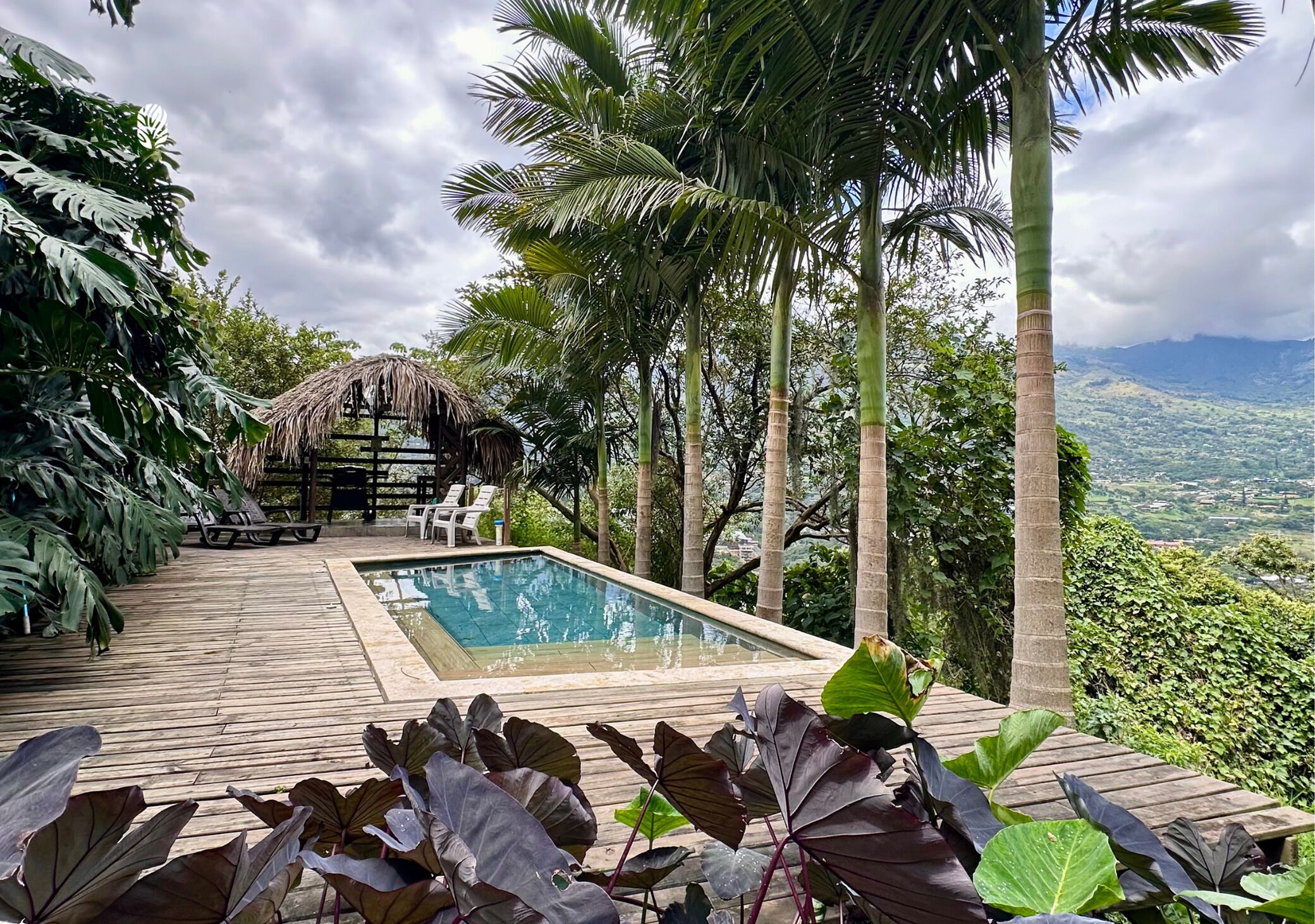 A pool area with a wooden deck, lounge chairs, and a thatched-roof structure.