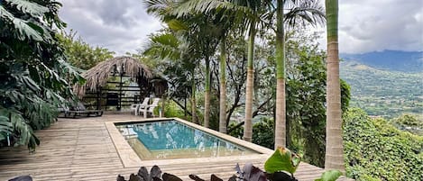 A pool area with a wooden deck, lounge chairs, and a thatched-roof structure.