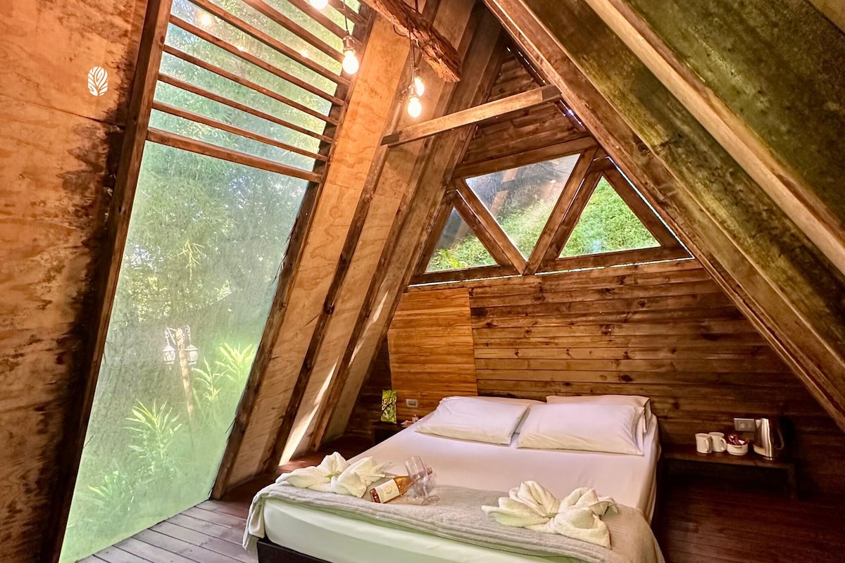 Een houten slaapkamer met een bed, kussens en een raam met uitzicht op groen.