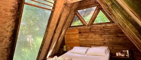A wooden cabin bedroom with a bed, pillows, and a window overlooking greenery.