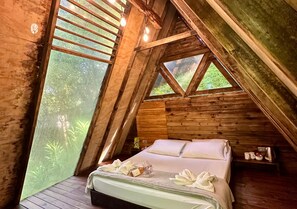 A wooden cabin bedroom with a bed, pillows, and a window overlooking greenery. - All Natural Glamping (Copacabana)