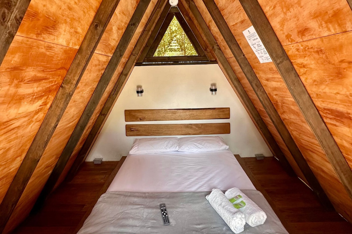Een enkel bed met een houten hoofdbord in een kamer met een driehoekige vorm, houten wanden en een raam aan de bovenkant.